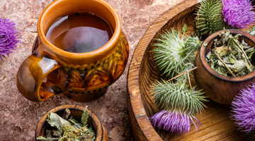 Herbal tea in a ceramic mug with dried milk thistle leaves and fresh purple milk thistle flowers on a wooden tray.