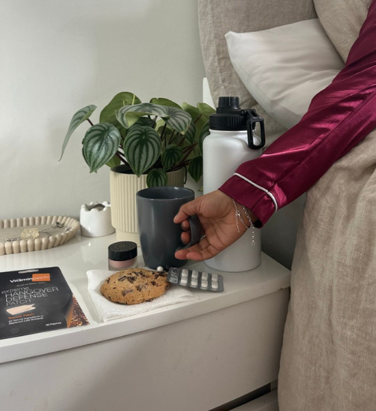 Person reaching for a mug on a bedside table with a cookie and Extreme Hangover Defense Patch.