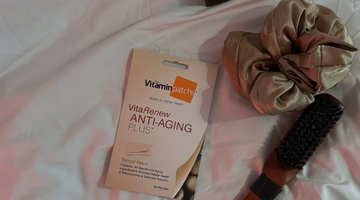 VitaRenew Anti-Aging Plus Patch pack placed on a bed beside a satin scrunchie and a wooden hairbrush.
