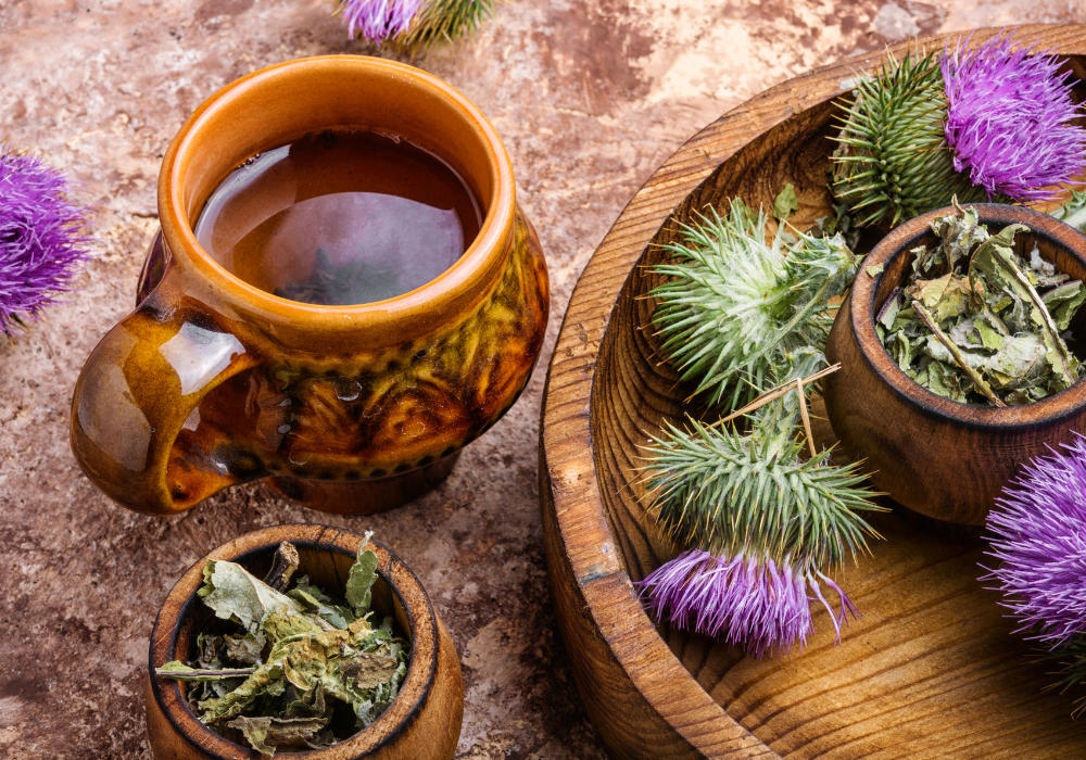 Herbal tea in a ceramic mug with dried milk thistle leaves and fresh purple milk thistle flowers on a wooden tray.