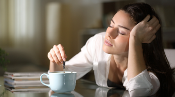 Woman looking tired while stirring coffee at a table indoors
