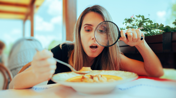 Woman using a magnifying glass to examine her food at an outdoor dining table.