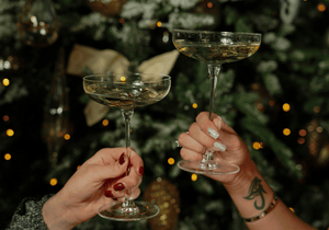 Two hands holding cocktail glasses in front of a Christmas tree.